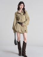 Woman wearing a khaki shirt dress, black studded belt, and brown boots against a neutral background.