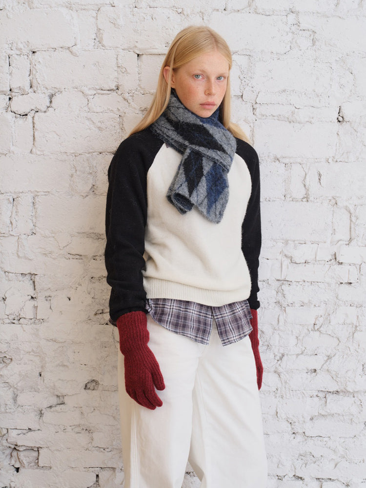 Close-up of a model wearing a cream and black raglan sleeve sweater, plaid shirt, white pants, and a blue argyle scarf.