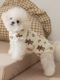 A Bichon Frise dog wearing a light yellow padded vest with a teddy bear print sits on a surface.