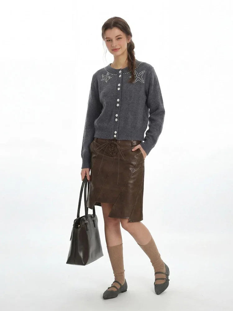 Full-body shot of a woman in a grey cardigan and brown skirt, holding a black handbag.