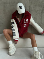 Woman in a white baseball cap and a red and white sherpa fleece bomber jacket, seated on a couch.