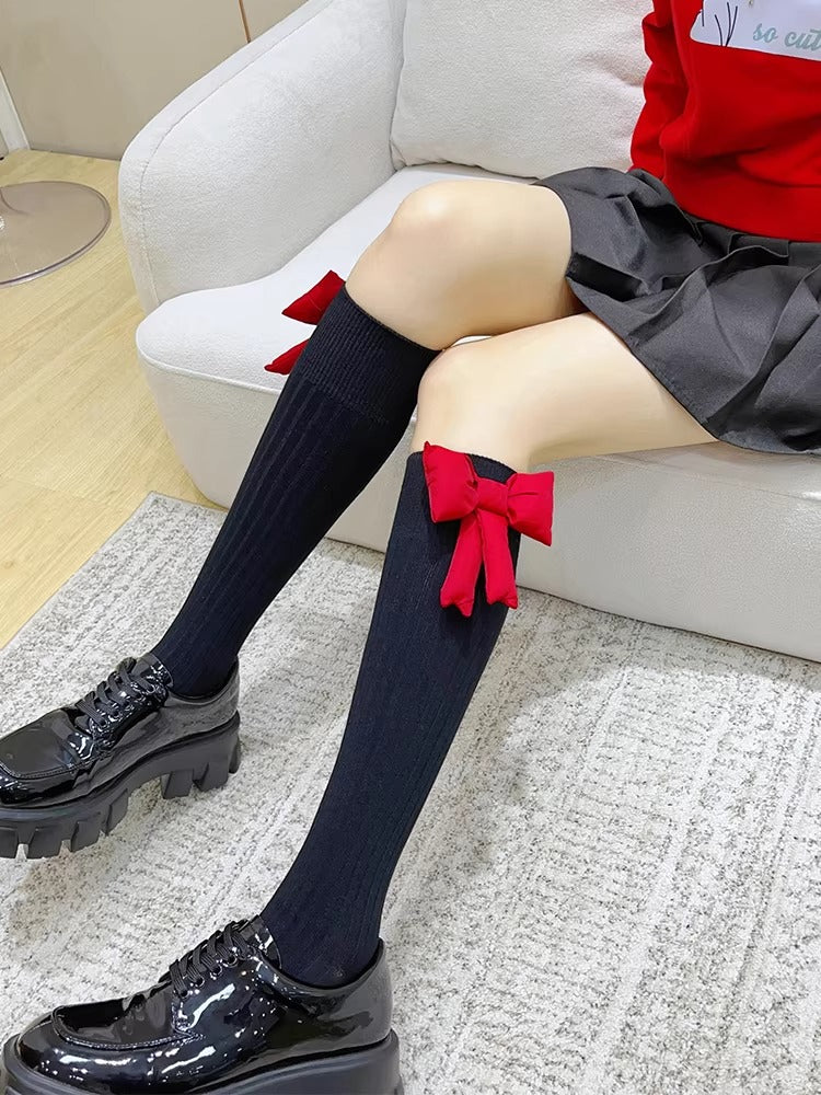 Legs of a person wearing black ribbed crew socks with red bow accents on the calf, paired with black patent platform shoes and a black pleated skirt.