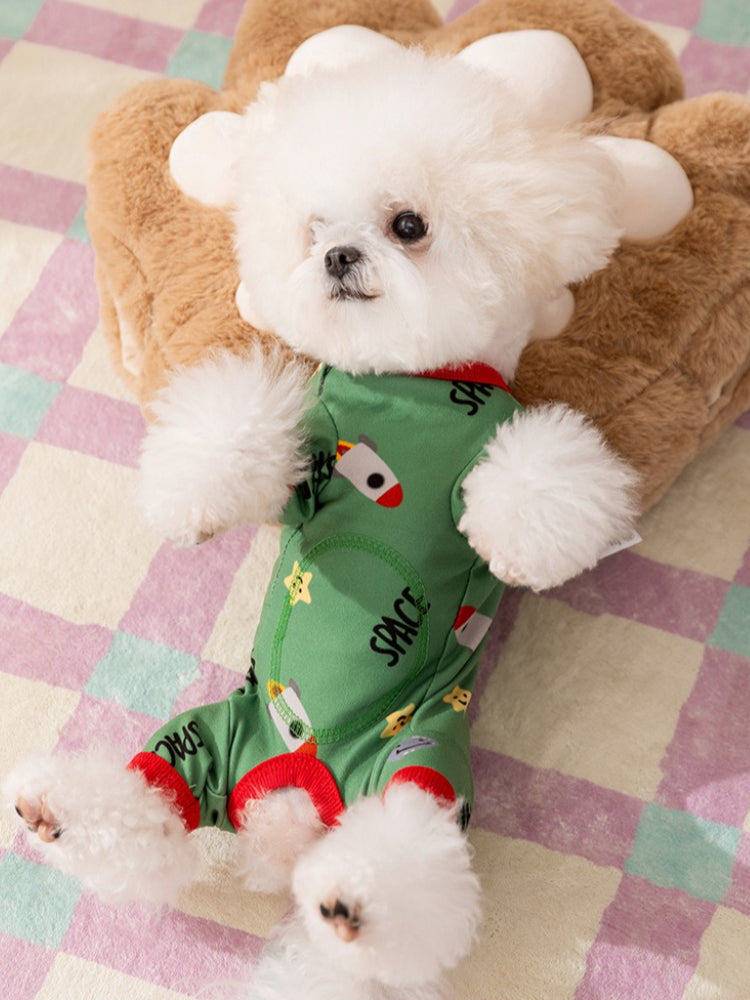 Small white dog wearing a green dog pajama with a space print and red trim