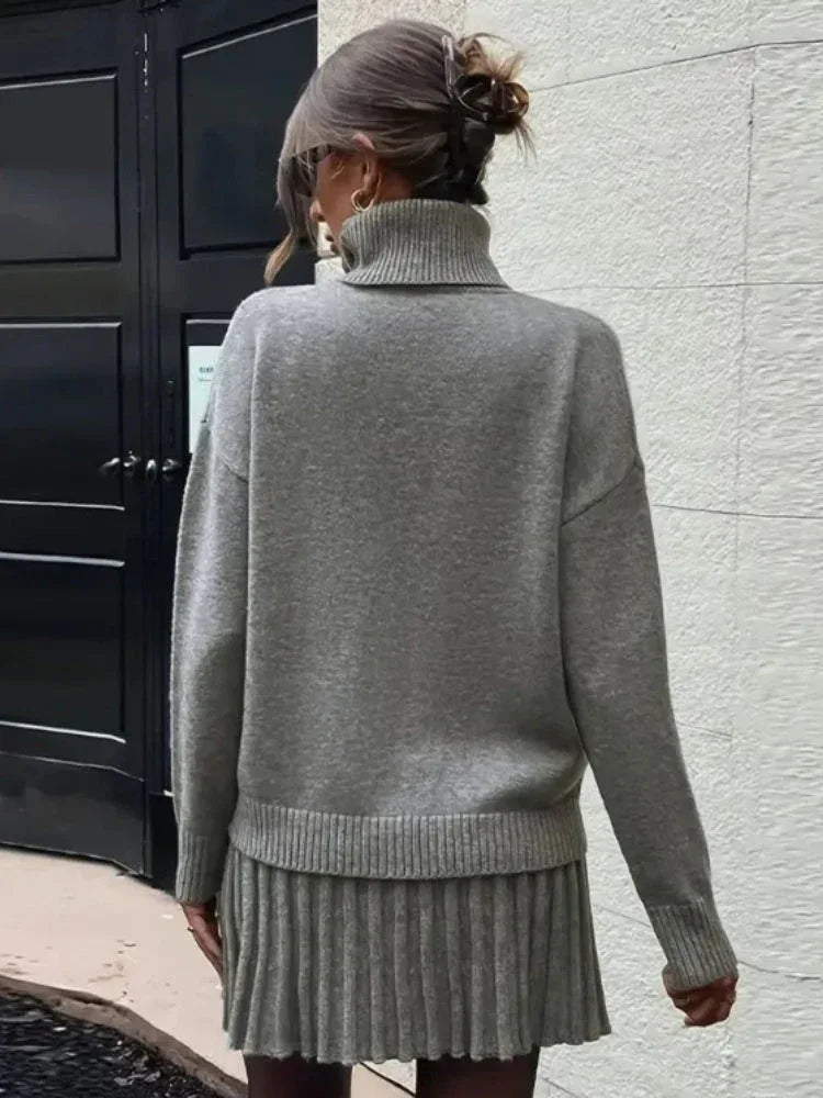 Woman wearing a grey knit turtleneck sweater and a matching grey pleated mini skirt, holding a black handbag.