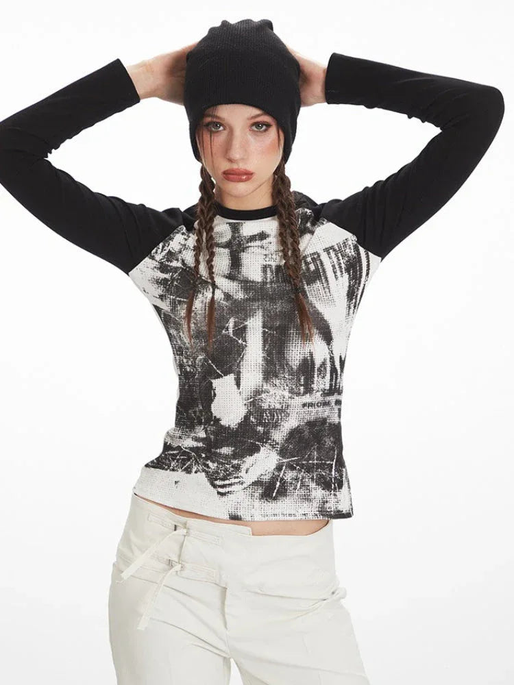 Woman posing with hands on head, wearing a black and white graphic print long sleeve top, white pants, and a black beanie