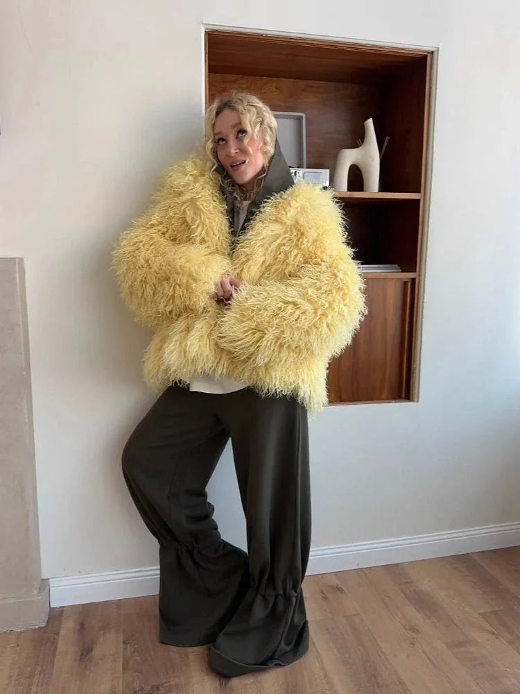 Woman wearing a yellow shaggy faux fur coat with dark green pants, leaning against a shelf.