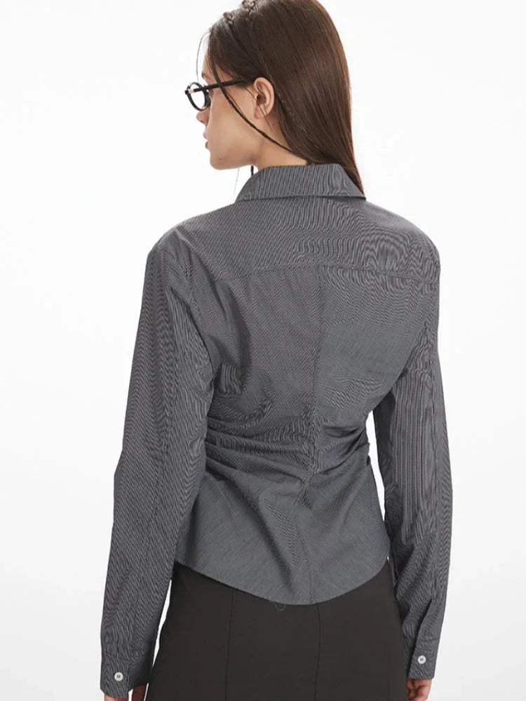 Rear view of a woman wearing a grey textured long-sleeve collared shirt with ruched waist detailing and a black midi skirt.