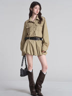 Full body shot of a woman in a khaki shirt dress with a black studded belt and knee-high boots.