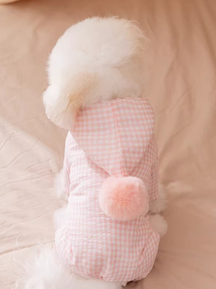 A Bichon Frise dog wearing a pink gingham hooded dog suit with a pink pom-pom accent.