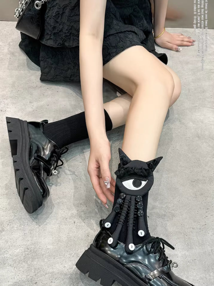 A person wearing black novelty socks with an eye applique and button details, and black platform shoes, sitting on the floor.