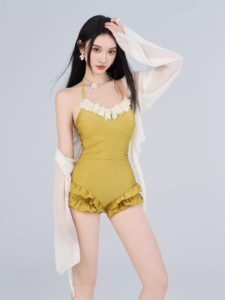 Woman wearing a mustard yellow one-piece swimsuit with floral trim and ruffled shorts, paired with a sheer white cover-up.