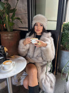 Woman wearing a long, light beige faux fur coat and a matching faux fur hat, sitting at a cafe table with coffee.