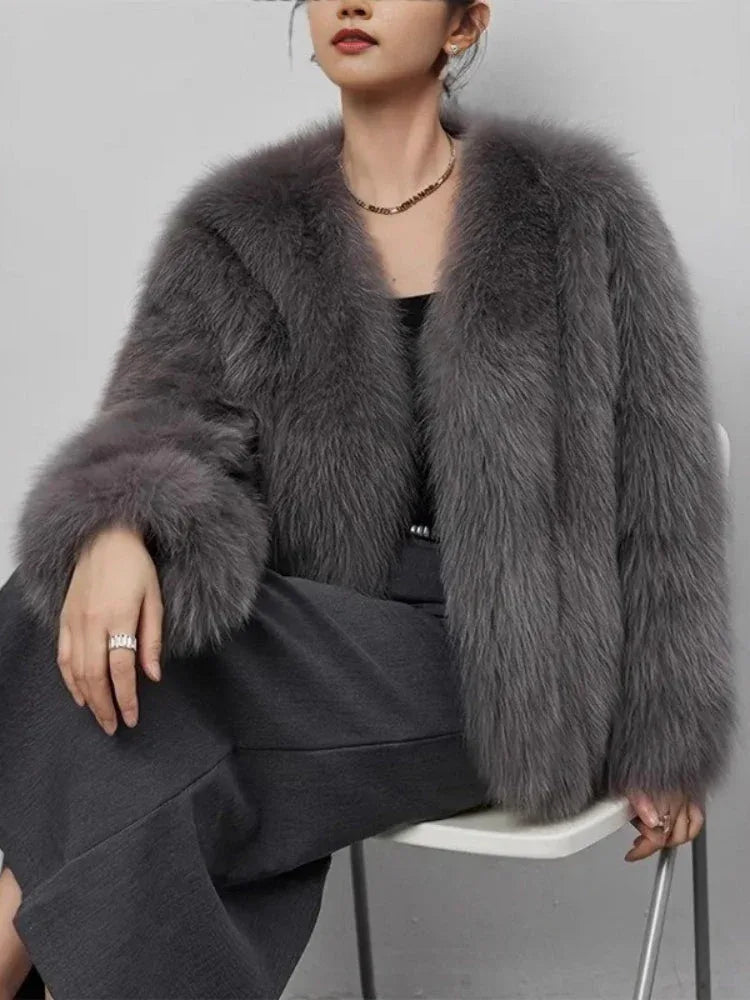 Woman sitting down wearing a grey faux fur jacket and dark trousers.
