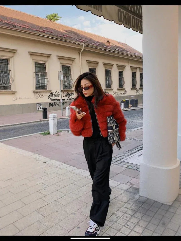 Woman wearing a red faux fur cropped jacket, black pants, and sneakers, walking and holding a phone.