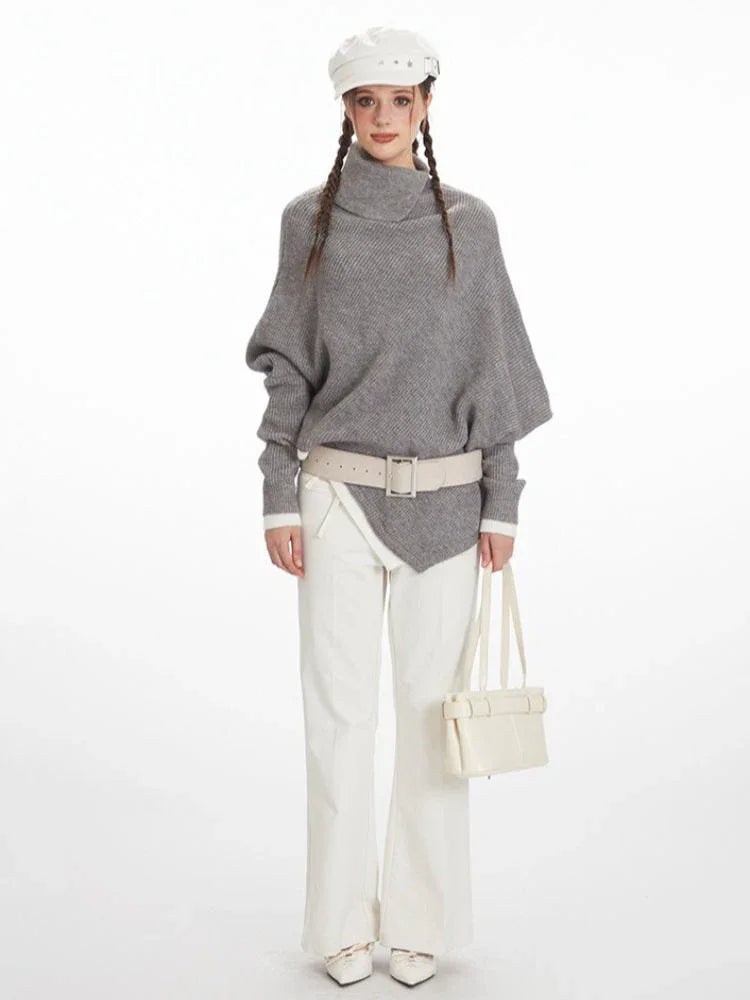Woman wearing a grey knit poncho with a turtleneck and batwing sleeves, white pants, a cream belt, and a white handbag. Main image