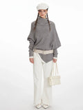Woman wearing a grey knit poncho with a turtleneck and batwing sleeves, white pants, a cream belt, and a white handbag.