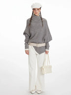 Woman wearing a grey knit poncho with a turtleneck and batwing sleeves, white pants, a cream belt, and a white handbag.