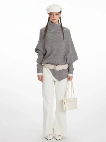 Woman wearing a grey knit poncho with a turtleneck and batwing sleeves, white pants, a cream belt, and a white handbag.