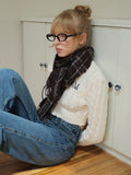 Woman leaning against a cabinet wearing a cream cable knit cropped sweater, plaid scarf, and blue jeans.
