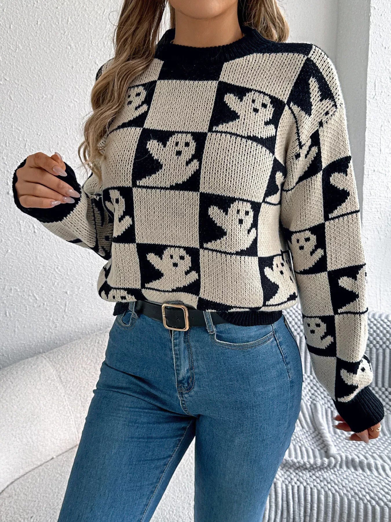 Close-up of a woman's torso wearing a black and cream checkerboard knit sweater with ghost print, blue jeans, and a black belt.