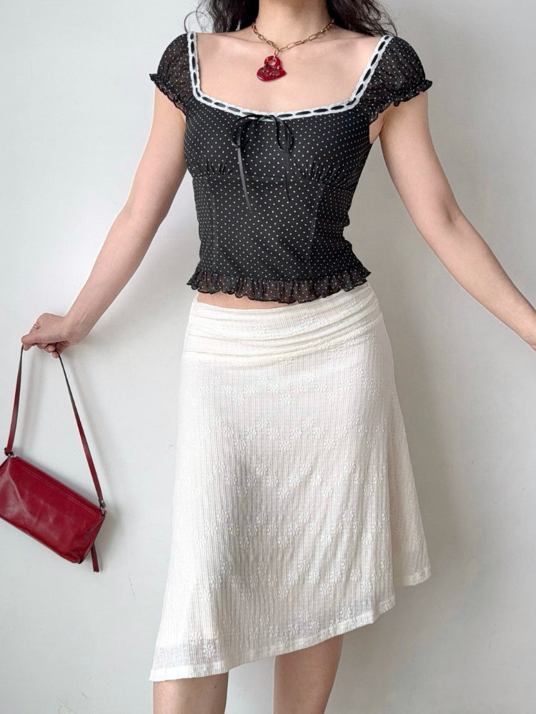 Full body shot of a woman in a black polka dot top and white asymmetrical skirt, holding a red handbag