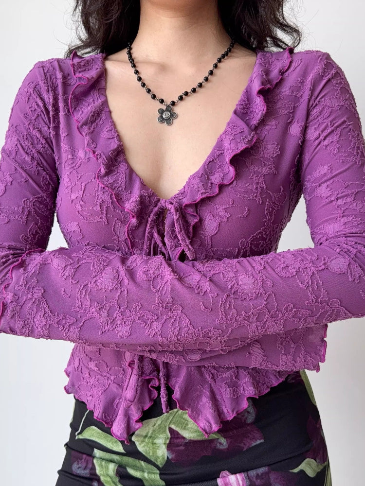 Close-up of a woman wearing a purple textured mesh long-sleeve top with a tie-front closure and ruffled V-neck.