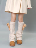Cream cable knit leg warmers with teddy bear embellishments and pom-poms, worn with a cream dress and tan boots.