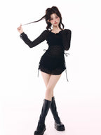 Woman posing in a black sheer long-sleeve top over a black bikini top and black ruffled hem shorts, with black platform boots.
