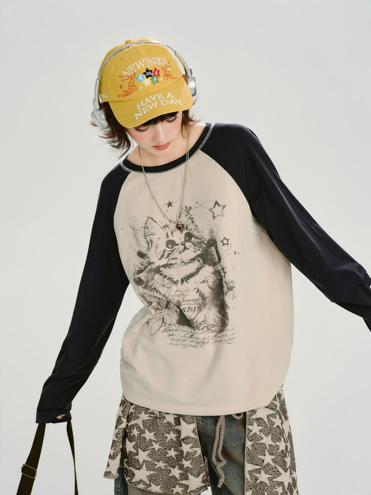 Woman wearing a cream long-sleeve raglan shirt with a graphic cat print and navy sleeves, paired with a patterned skirt and jeans.