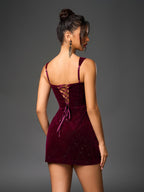 Woman in a short, deep red velvet dress with spaghetti straps and sparkling embellishments, back view showing lace-up detail.