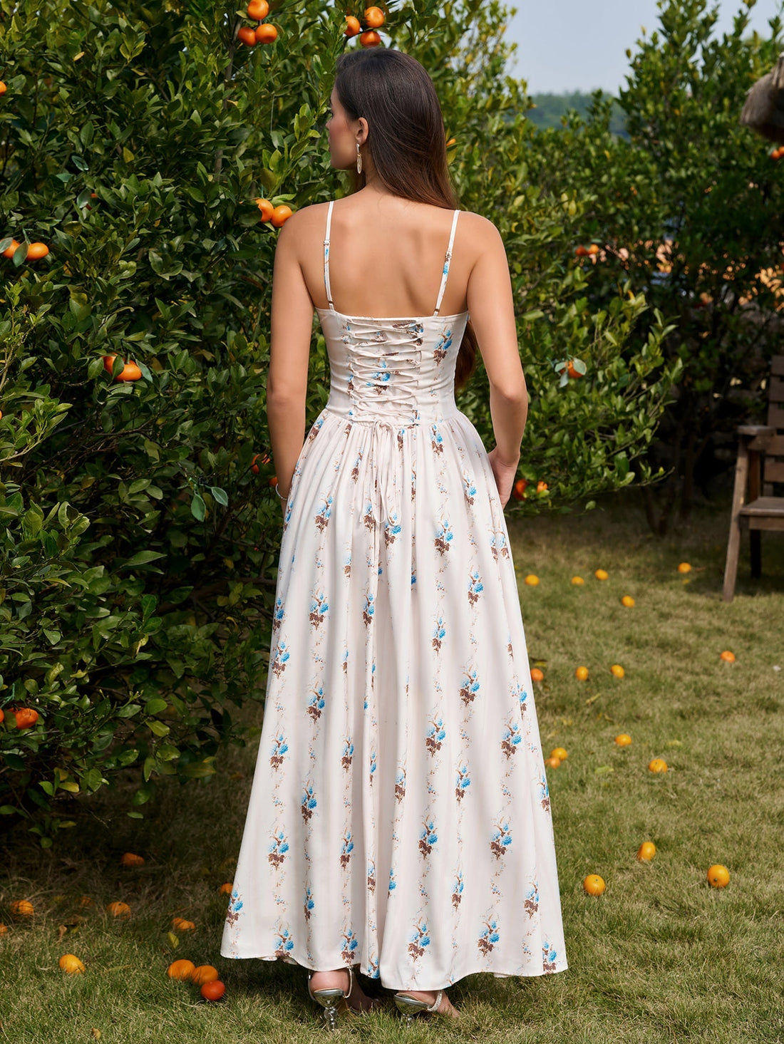 Woman wearing a white floral print maxi dress with spaghetti straps and a square neckline, standing in an orchard.