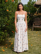 Woman wearing a white floral print maxi dress with spaghetti straps and a square neckline, standing in an orchard.