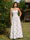 Woman in a white floral print maxi dress with spaghetti straps and side pockets, posing with hand on hip in a garden.