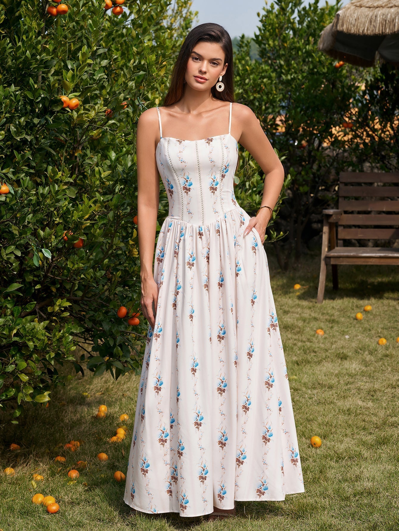 Woman in a white floral print maxi dress with spaghetti straps and side pockets, posing with hand on hip in a garden.