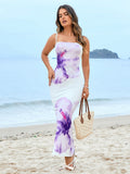Woman in a white bodycon maxi dress with a purple floral print and spaghetti straps, holding a straw handbag on a beach.