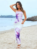 Woman in a white bodycon maxi dress with a purple floral print and spaghetti straps, with hand on hip on a beach.