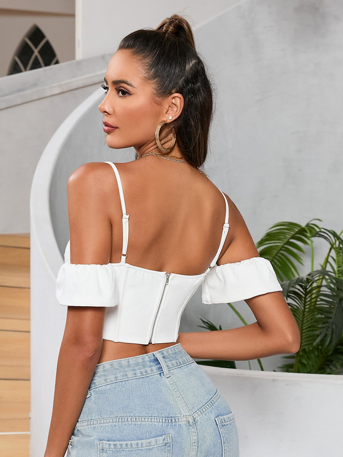 Woman wearing a white off-the-shoulder corset crop top with spaghetti straps and ruffled sleeves, paired with light wash denim jeans.
