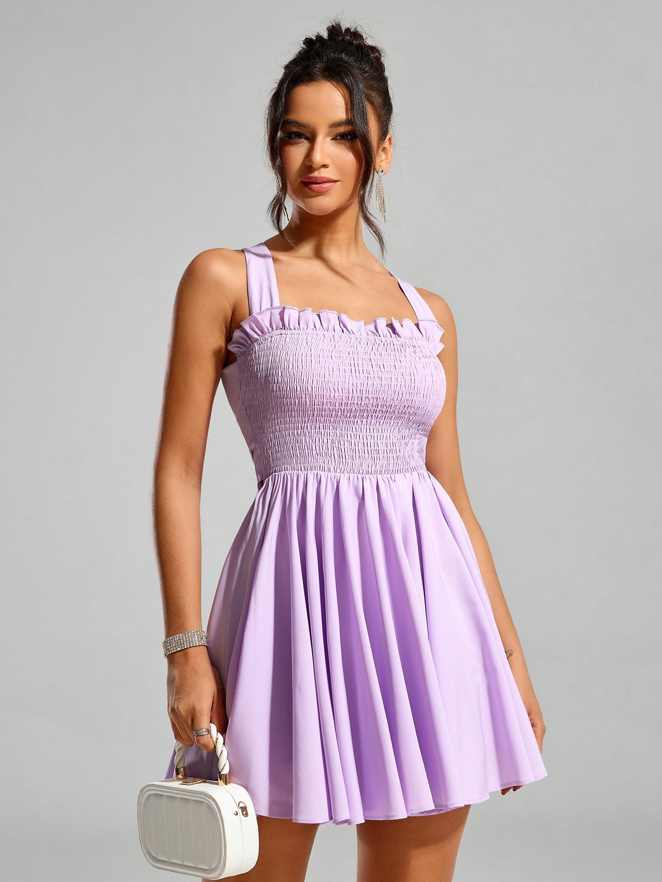 Front three-quarter view of a woman in a lavender smocked mini dress holding a white handbag