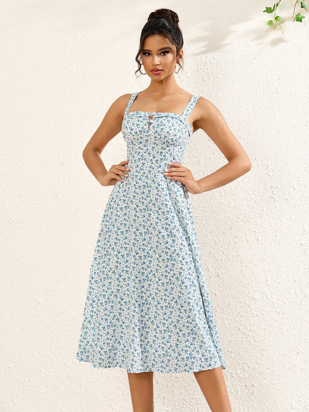 Woman with hands on hips wearing a white midi dress with a blue floral print and tie detail at the bust.