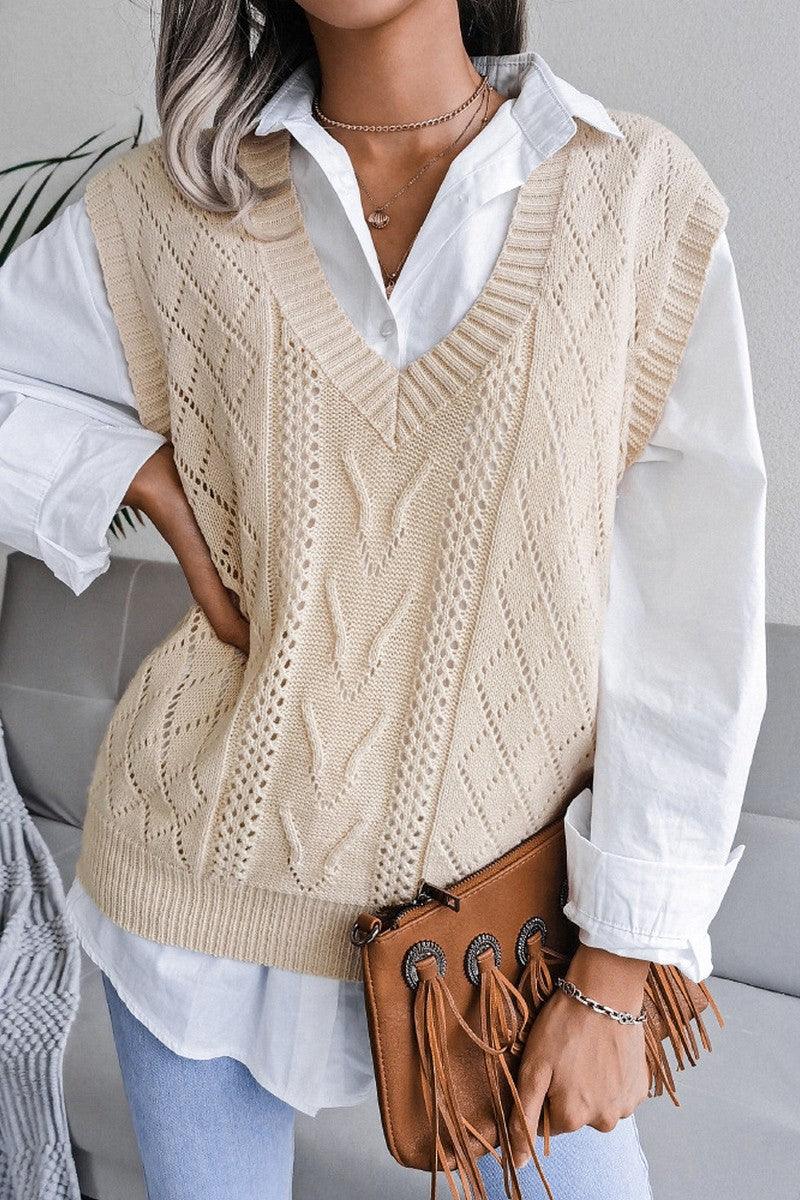 Front view of a woman wearing a beige cable knit V-neck sleeveless sweater vest over a white collared shirt, holding a brown fringed clutch.