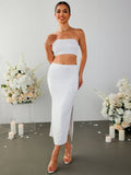 Woman wearing a white strapless cropped top and a white midi skirt with a side slit, standing in front of floral arrangements.