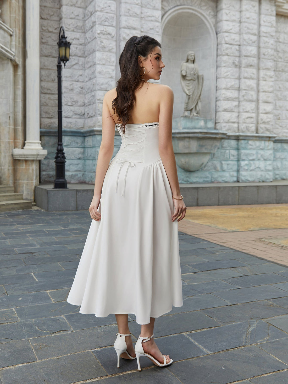 Woman wearing a white strapless midi dress with black tie detail, leaning against a lamppost