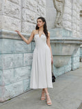 Woman posing in a white halter midi dress with contrast trim, leaning against a stone wall