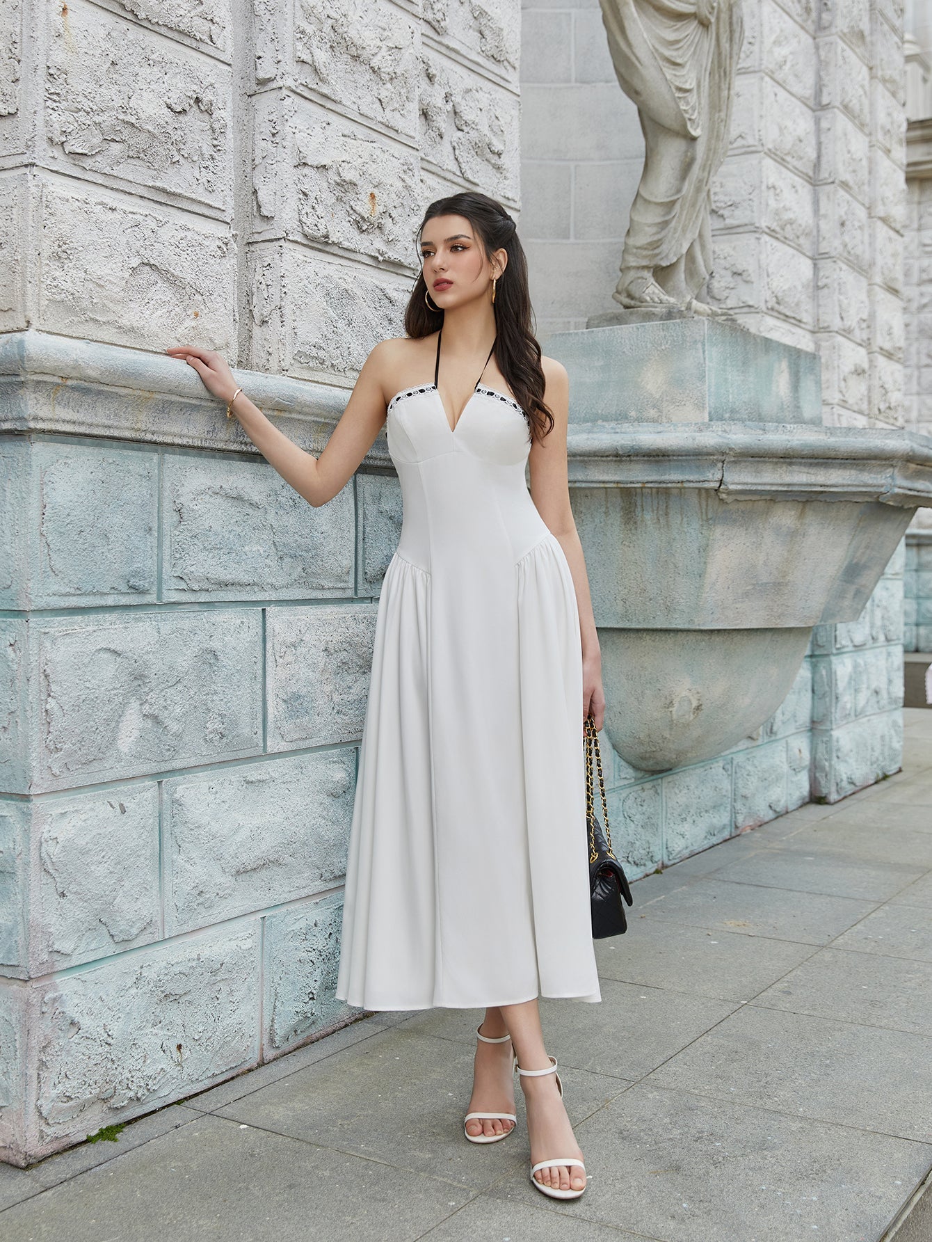 Woman posing in a white halter midi dress with contrast trim, leaning against a stone wall