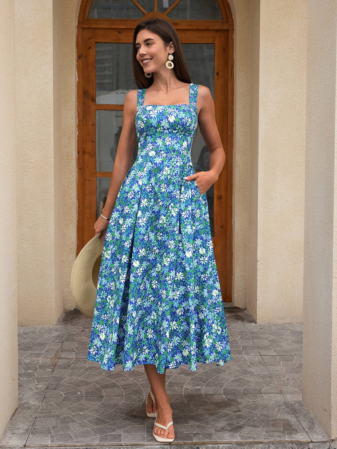 Woman wearing a blue floral print midi dress with a square neckline and wide straps, holding a straw hat.
