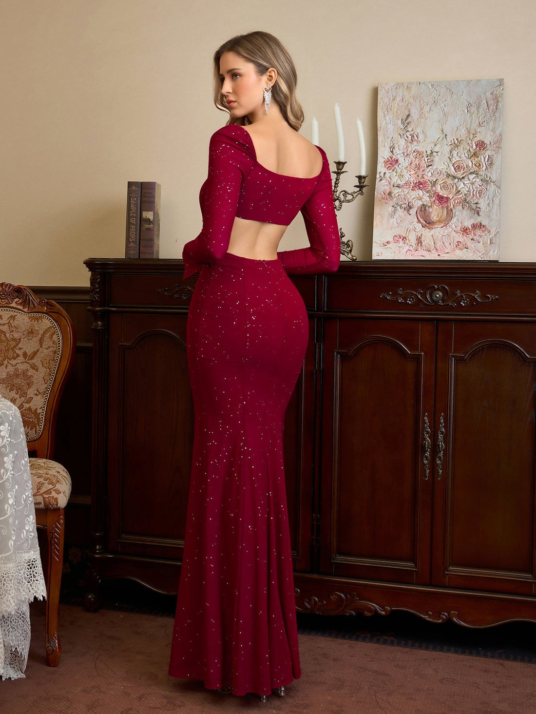 Woman in a long-sleeved, burgundy, sequined gown with a square neckline and a high slit, standing in a room.