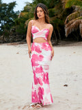 Woman standing on a beach wearing a pink and white tie-dye maxi dress with cut-out detail and palm trees in the background.