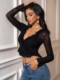 Woman leaning against a wall, wearing a black long-sleeve sheer lace cropped top with ruffled trim and deep V-neck, paired with blue jeans.