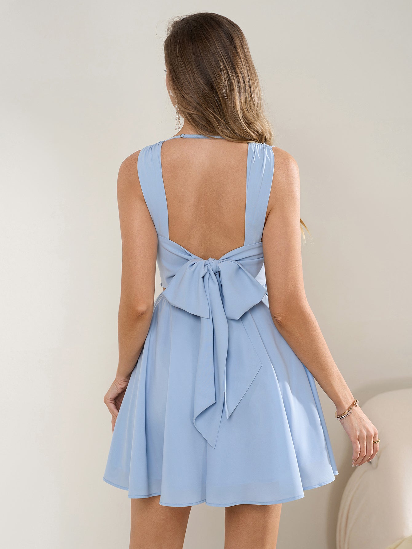 Back view of a woman in a light blue sleeveless cowl neck fit and flare dress with an open back and bow detail.