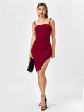 Woman wearing a red embellished ruched midi dress with spaghetti straps and an asymmetrical hem, holding a black clutch.